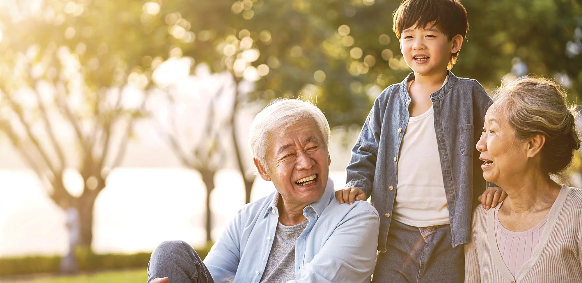 Photo of grandson, grandfather and grandmother sitting chatting on grass outdoors in park at dusk