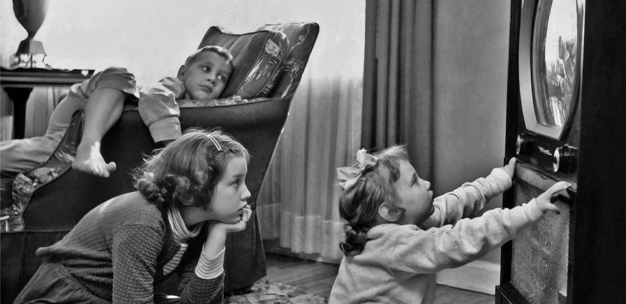 Photo de trois enfants regardant la télévision. L’un d’eux est assis tout près du téléviseur vers les années 1960.