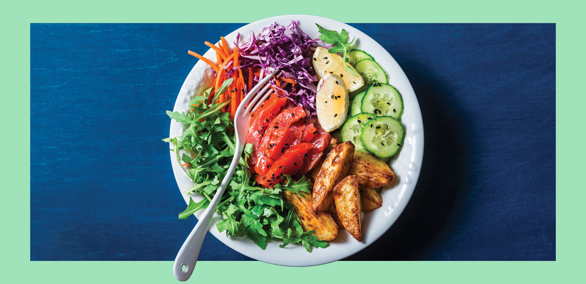 Photo of Smoked salmon, baked potatoes, vegetables buddha power bowl on blue background, top view. Red cabbage, carrots, arugula, potatoes, smoked salmon fish bowl.